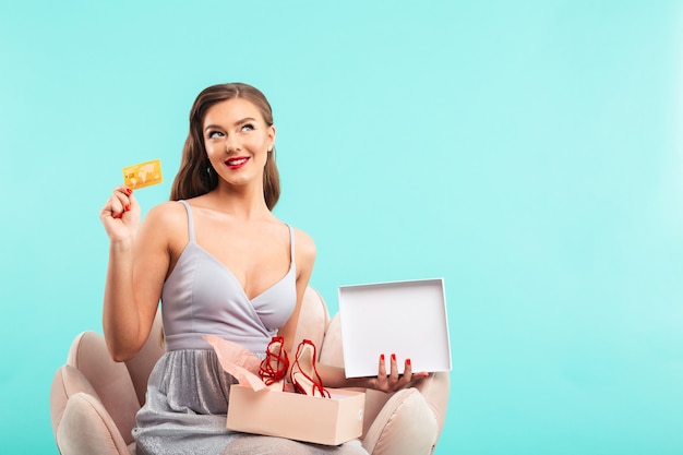 Mujer de compras de 20 años en vestido sosteniendo la caja actual con zapatos y tarjeta de crédito mientras está sentado en un sillón, aislado sobre una pared azul