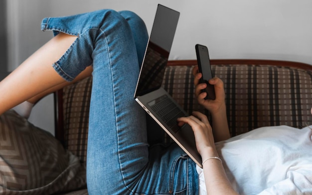 Una mujer con una computadora portátil está acostada en un sofá y usa un teléfono inteligente