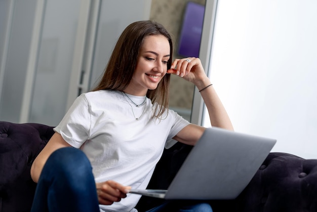 Mujer con computadora portátil de rodillas Mujer sentada con las piernas cruzadas en el sofá Trabajando en la computadora portátil desde casa