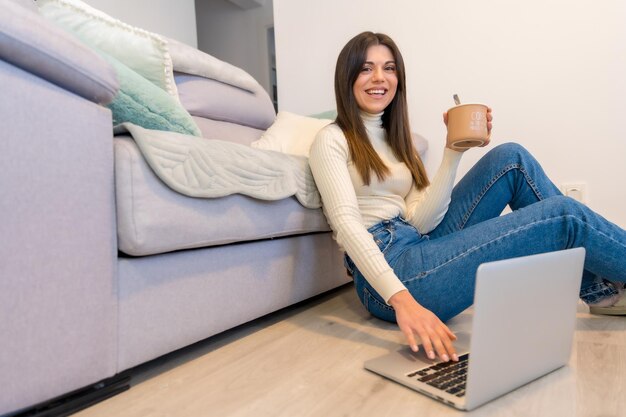 Mujer con una computadora sentada debajo del sofá con un café caliente Blog de redes sociales del milenio