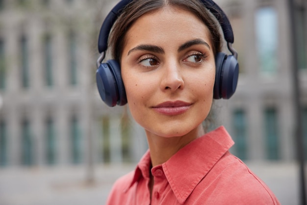 Foto mujer concentrada en algún lugar pasea al aire libre en la ciudad al aire libre lleva auriculares estéreo inalámbricos en los oídos escucha pistas de audio o podcast durante el ocio