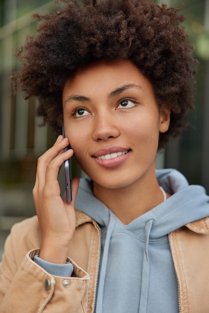mujer concentrada en algún lugar tiene conversación telefónica mantiene el teléfono inteligente cerca de la oreja usa sudadera con capucha y chaqueta plantea conversaciones en itinerancia