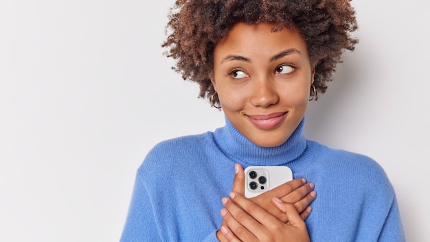 Mujer concentrada a un lado pensativamente tiene expresión soñadora sostiene el teléfono móvil cerca del cuerpo contento de recibir un mensaje de su novio aislado en blanco