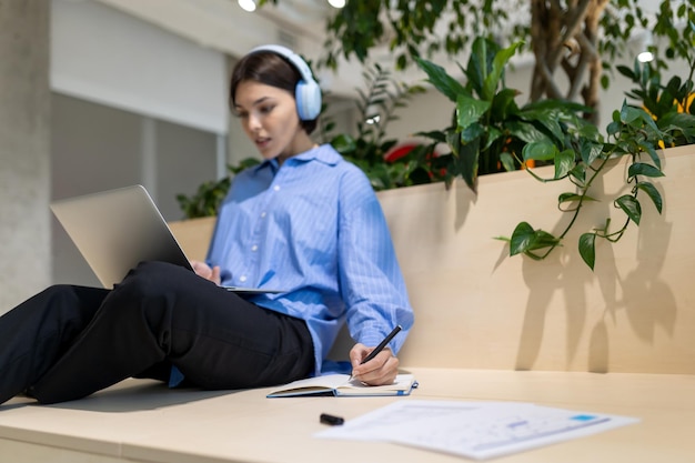 Una mujer concentrada y seria con auriculares mirando el monitor de la computadora y tomando notas en el cuaderno