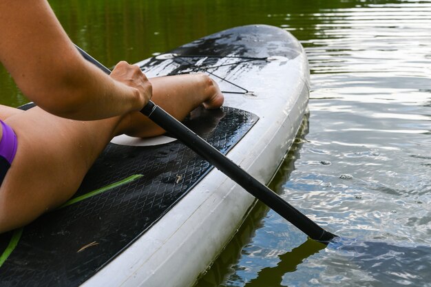 Una mujer conduce en el Sup Board a través de un canal estrecho rodeado de hierba densa. Vacaciones de fin de semana activo naturaleza salvaje al aire libre. Una mujer está sentada con las piernas estiradas.