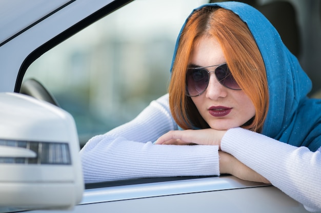 Mujer conductora en bufanda y gafas de sol mirando por la ventana delantera del coche.