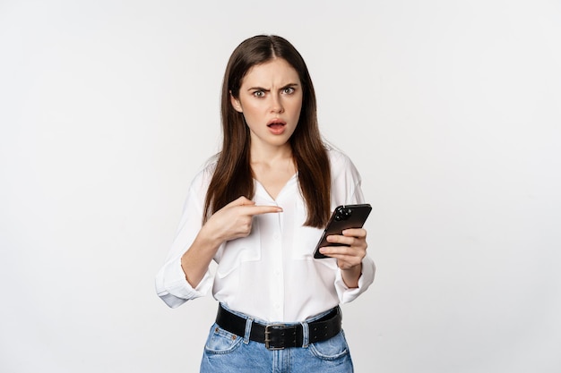 Mujer confundida apuntando al teléfono móvil y luciendo desconcertada, no puede descifrar algo, de pie sobre fondo blanco.