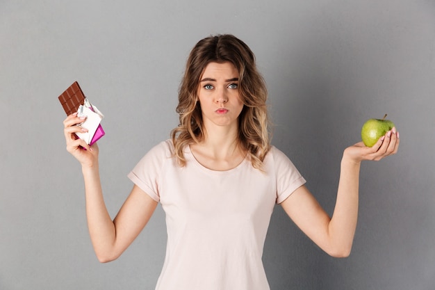 Mujer confundida en camiseta con chocolate y manzana fresca mientras sobre gris