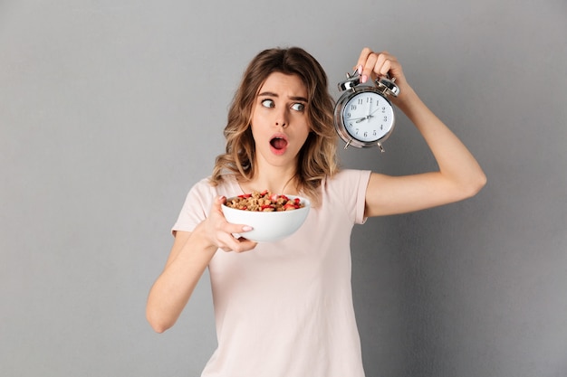 Mujer confundida en camiseta con comida sana en plato y despertador sobre gris