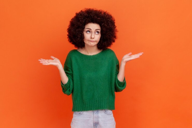 Foto mujer confundida con peinado afro con suéter verde de estilo casual extendiendo las manos a un lado no sé qué hacer mirando hacia otro lado toma de estudio interior aislada en fondo naranja