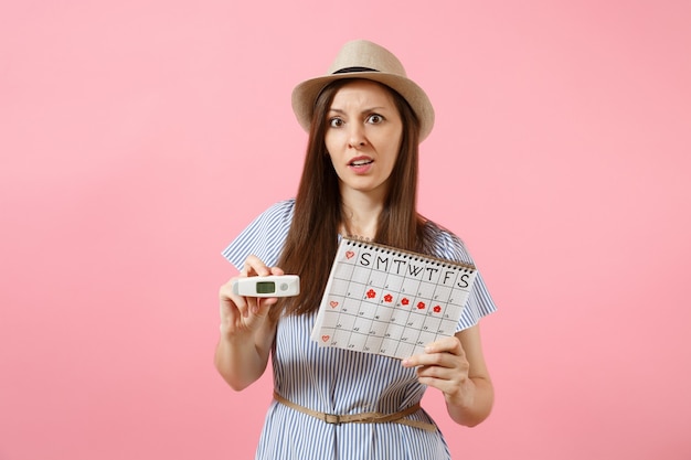 Mujer confundida en vestido mantenga en la mano el termómetro, calendario de períodos femeninos para comprobar los días de menstruación aislados sobre fondo rosa. Atención médica, concepto ginecológico de ovulación. Copie el espacio.