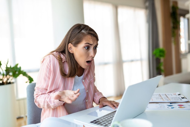 Mujer conmocionada agotada por trabajar, estudiar duro, aburrida y frustrada mirando la computadora portátil, la cabeza descansando en la mano, espacio brillante, grandes ventanas, concepto de estrés en el hogar.