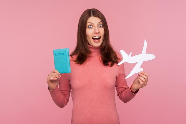 Mujer conmocionada con cabello castaño en suéter rosa sosteniendo en las manos un avión de papel y pasaporte, preparándose para un viaje repentino, asombrada con la aventura. Disparo de estudio interior aislado sobre fondo rosa