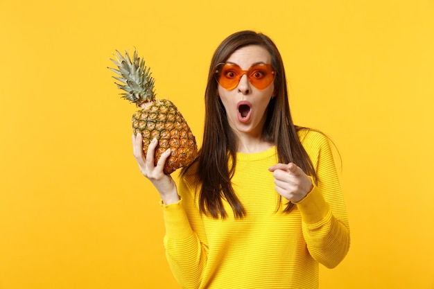 Mujer conmocionada con gafas de corazón manteniendo la boca abierta apuntando con el dedo índice a la cámara, sostenga fruta fresca de piña madura aislada en un fondo amarillo anaranjado. Estilo de vida vivo de la gente, concepto de vacaciones relajantes.