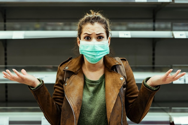 Foto mujer conmocionada con máscara protectora gesticulando mientras está de pie frente a un estante vacío en el supermercado durante la pandemia del coronavirus