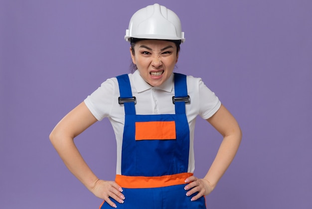 Mujer constructora asiática joven molesta con casco de seguridad blanco poniendo las manos en la cintura y mirando