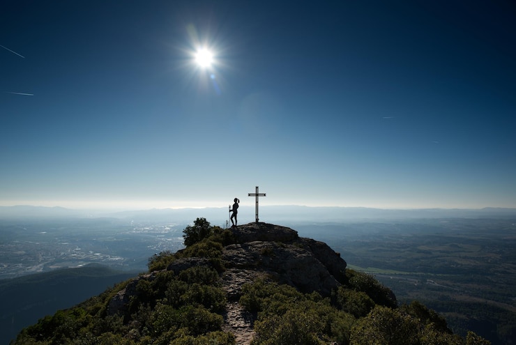 Foto mujer contemplando un paisaje retroiluminado desde la cima de una colina donde hay una cruz