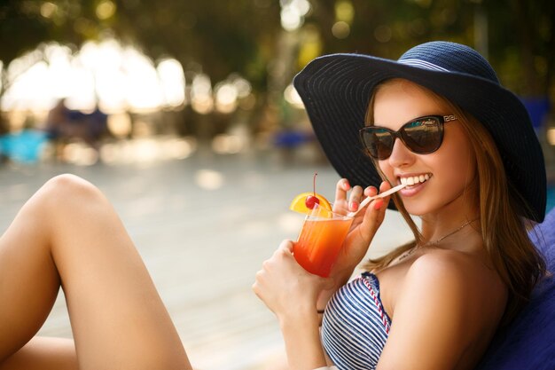 Mujer con copa de cóctel relajándose bajo el sol tropical cerca de la piscina en una tumbona