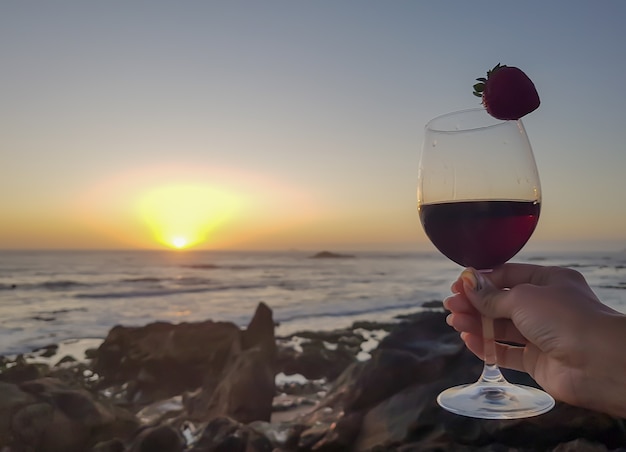 Mujer con copa de vino tinto y fresa en la playa. Puesta de sol maravillosa