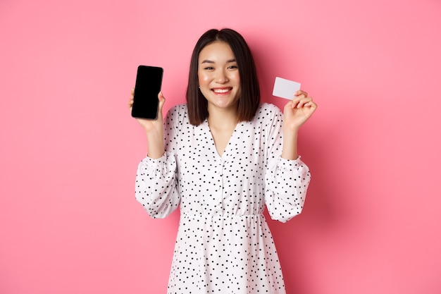 Mujer coreana alegre que muestra la pantalla del teléfono inteligente y la tarjeta de crédito, pagando el pedido por Internet