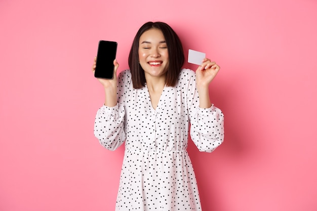 Mujer coreana alegre que muestra la pantalla del teléfono inteligente y la tarjeta de crédito que paga para la demostración del pedido por internet ...