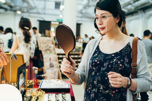 Mujer coreana asiática embarazada de moda joven probándose gafas nuevas en un vendedor de tiendas ópticas en un mercado creativo. Hermosa niña de maternidad usando anteojos y mirándose en el espejo. futura mamá de compras