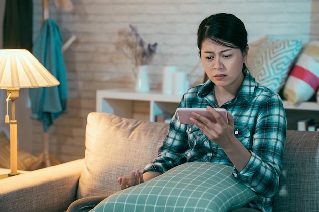 mujer coreana asiática molesta viendo contenido en línea en un teléfono inteligente sentado en un sofá en una sala de estar oscura en casa por la noche. una chica triste se emborracha viendo una película de amor, una película romántica y miserable en el teléfono celular, frunciendo el ceño.