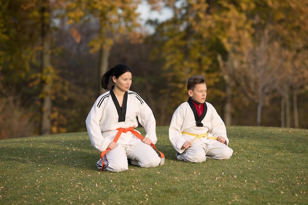 Mujer coreana del sofá y un niño en kimoni sentados en la hierba verde Mujer enseñando taekwondo a un niño pequeño en el parque de otoño