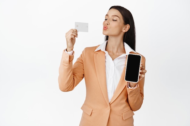 Mujer corporativa besando su tarjeta de crédito que muestra el pedido en la pantalla vacía de la pantalla del teléfono móvil en el teléfono inteligente de pie en traje sobre fondo blanco