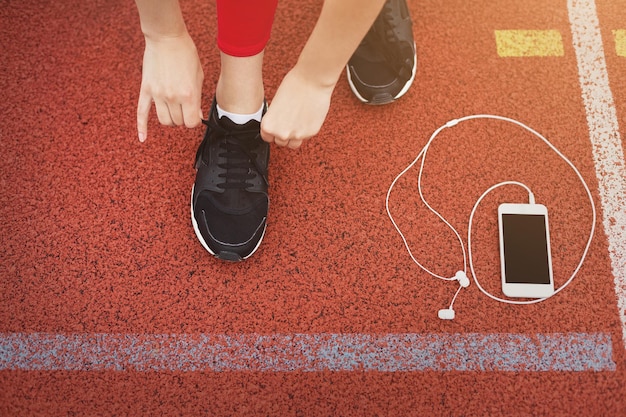 Mujer corredor atar cordones de zapatos en pista de atletismo con smartphone y auriculares