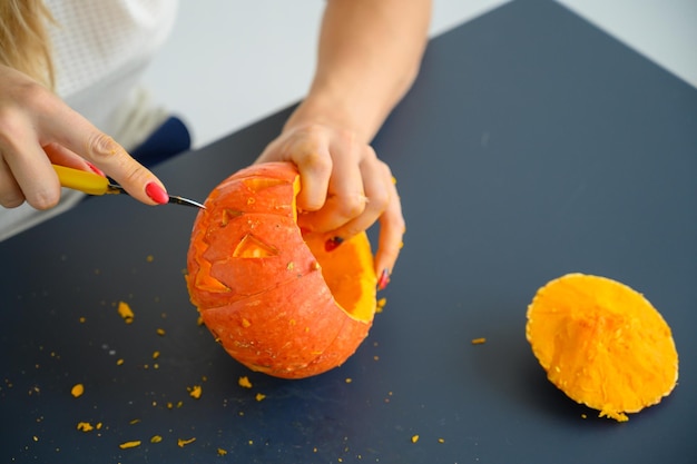 Una mujer corta una cara con un cuchillo de papelería en una calabaza para lámparas de Halloween en una mesa negra Primer plano de manos femeninas haciendo jacko39lantern en la víspera de todos los santos Vista frontal