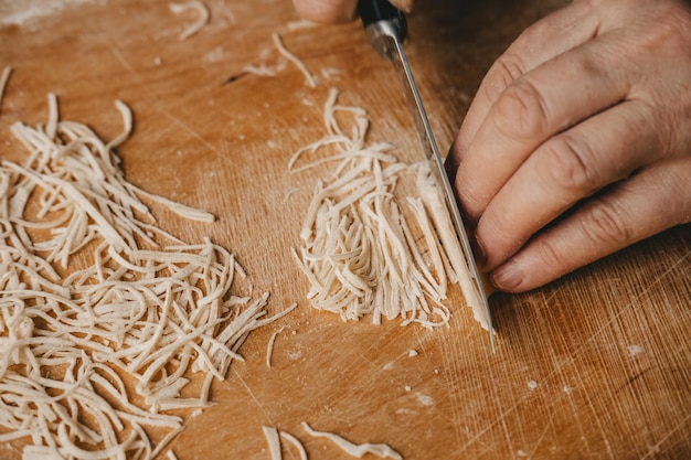 Una mujer corta la masa con un cuchillo y hace fideos caseros en la cocinaPasta hecha en casa por las manos de una mujer