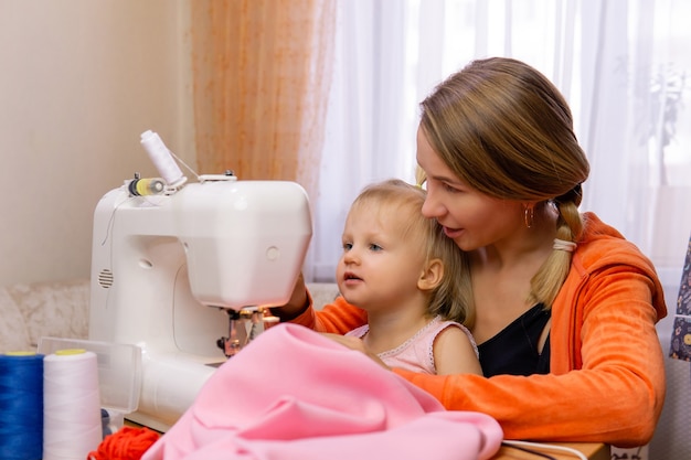 Mujer cose en casa con una máquina de coser eléctrica y se relaciona con su hija pequeña