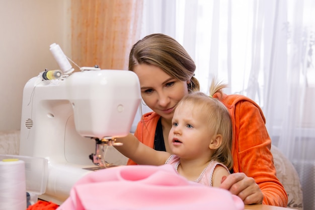 Mujer cose en casa con una máquina de coser eléctrica y se relaciona con su hija pequeña