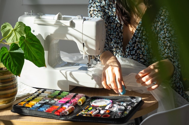 Una mujer cose tul en una máquina de coser eléctrica en un interior blanco y moderno de una casa con grandes ventanas plantas de interior Comodidad en la casa el pasatiempo de un ama de casa