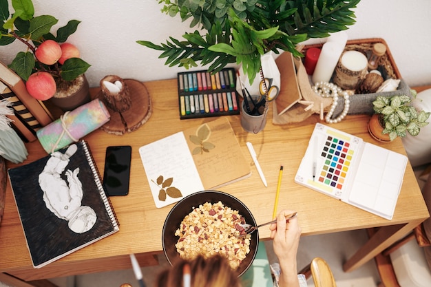 Mujer creativa comiendo muesli con bayas cuando está sentada en su escritorio con dibujos, diarios y pinturas, vista desde la parte superior