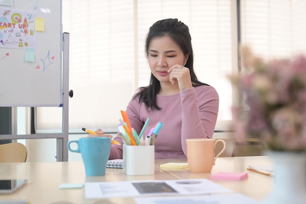 Mujer creativa joven concentrada sentada en su escritorio de oficina y tomando notas de datos importantes