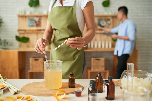 Mujer creativa mezclando una base de jabón derretido con una varilla de vidrio al agregar unas gotas de aceite esencial