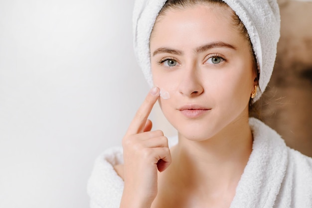 Mujer con crema facial en albornoz con una toalla en la cabeza después de ducharse en casa. aplicar crema para la piel sobre la piel.