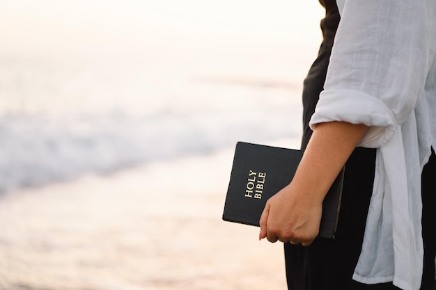 Mujer cristiana tiene la Biblia en sus manos leyendo la Santa Biblia en el mar durante la hermosa puesta de sol