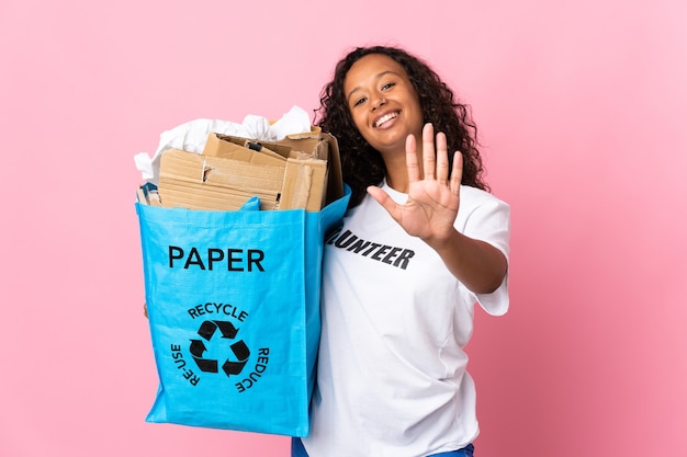 Mujer cubana sosteniendo una bolsa de reciclaje llena de papel para reciclar aislado en rosa contando cinco con los dedos