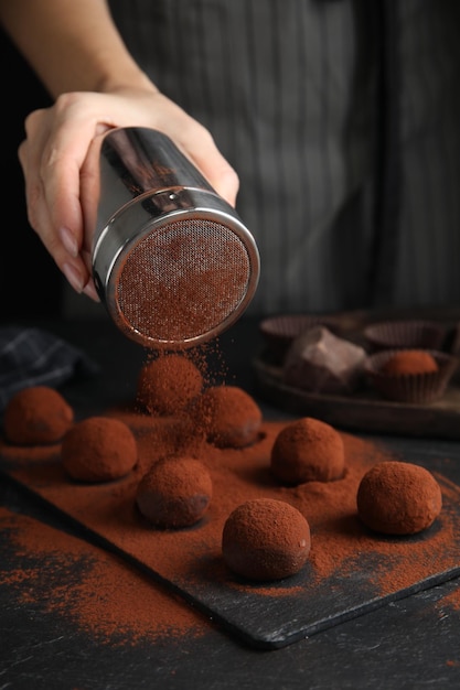 Mujer cubriendo deliciosas trufas de chocolate con cacao en polvo en el primer plano de la mesa negra