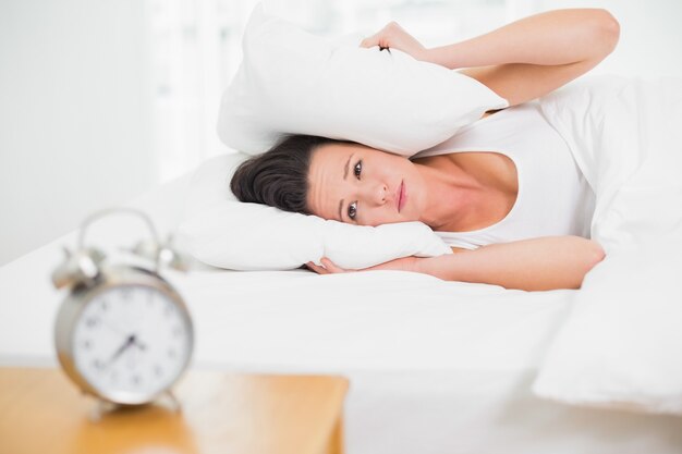 Mujer cubriendo las orejas con almohada y despertador en la mesa auxiliar