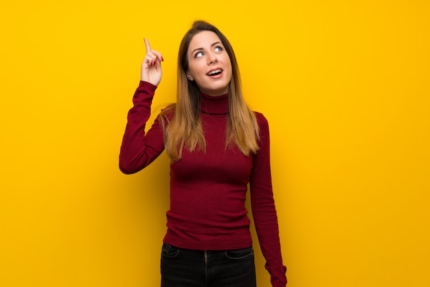 Mujer con cuello alto sobre una pared amarilla con la intención de realizar la solución mientras levanta un dedo.