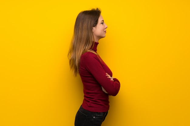 Mujer con cuello alto sobre pared amarilla en posición lateral