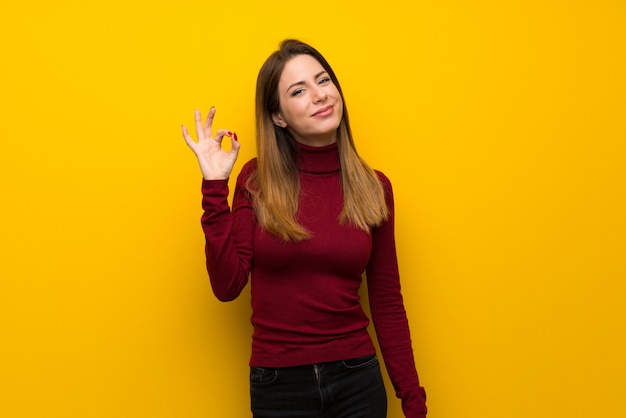 Mujer con cuello alto sobre una pared amarilla que muestra el signo correcto con los dedos