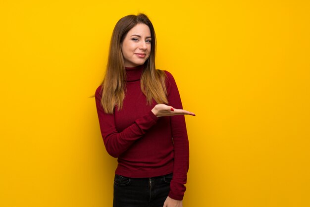 Mujer con cuello alto sobre una pared amarilla que presenta una idea mientras mira sonriendo hacia