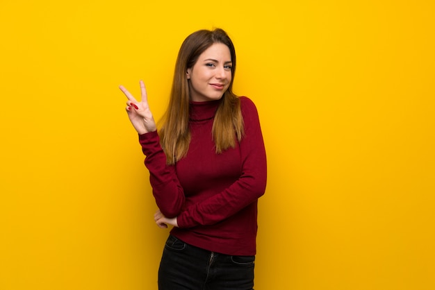 Mujer con cuello alto sobre pared amarilla sonriendo y mostrando el signo de la victoria