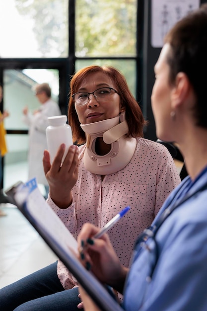 Mujer con cuello cervical sosteniendo una botella de pastillas mientras la enfermera escribe el tratamiento con medicamentos en el informe durante el examen. Paciente lesionado con cita en la sala de espera del hospital