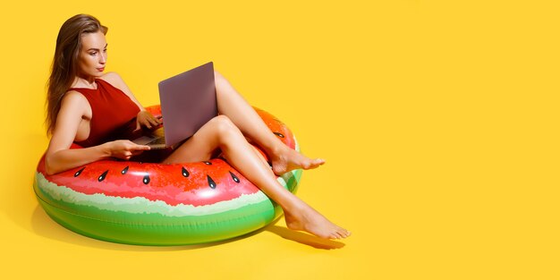 La mujer de cuerpo entero en bikini rojo se sienta en el anillo inflable de la sandía del baño utiliza la PC portátil aislada en el fondo amarillo. Viaje de pasajeros al extranjero escapada de fin de semana. Concepto de viaje de vuelo aéreo.